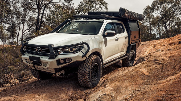 A white Mazda BT-50 equipped with an off-road bull bar and rugged tires, navigating a rocky trail in a wooded area. The vehicle is fitted with a roof rack and additional storage, showcasing its readiness for adventurous expeditions.