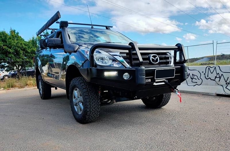 A silver Mazda BT-50 featuring a heavy-duty bull bar, an awning, and roof rack, parked on a paved surface near a fenced area. The vehicle is prepared for outdoor activities, combining both functionality and style.